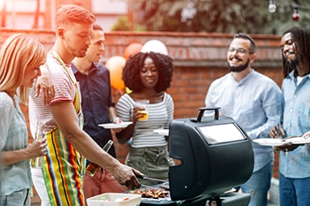 Group of young people having fun at barbecue party in a backyard. Selective focus to man grilling meats to his friends. Blonde woman holding a bottle in her hand. They are enjoy spending time together.