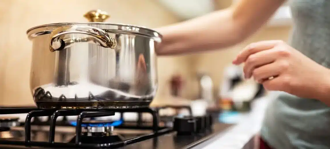 A woman in a blue shirt cooks with a saucepan on a gas stove.