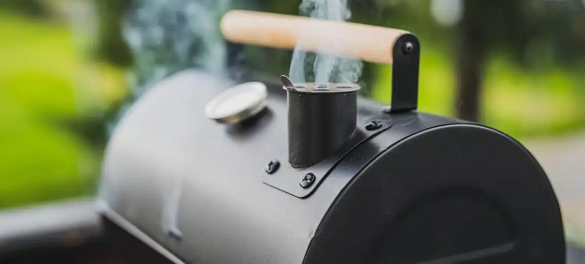 Smoke coming out of a smokestack of a small black smoker grill or barbecue on a green background.